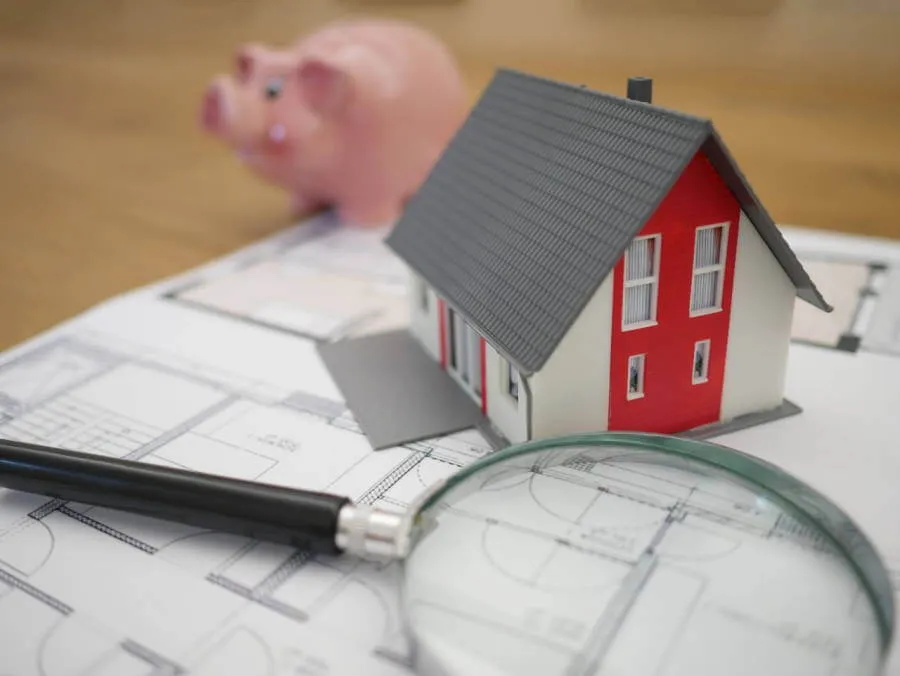 Red toy house on building plans with a piggy bank in the background