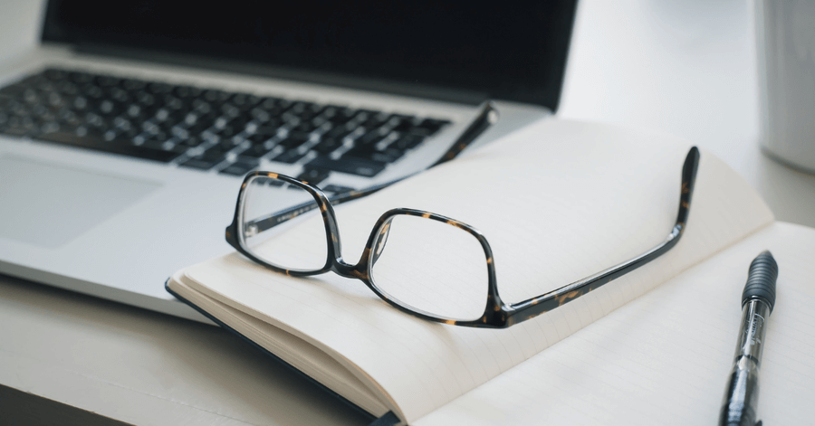 Glasses rested on top of an open notebook with a laptop open in the background