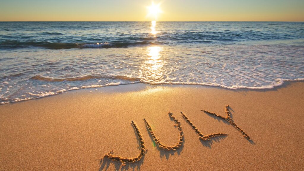 image of the word july written in the sand against a sea and sunset backdrop
